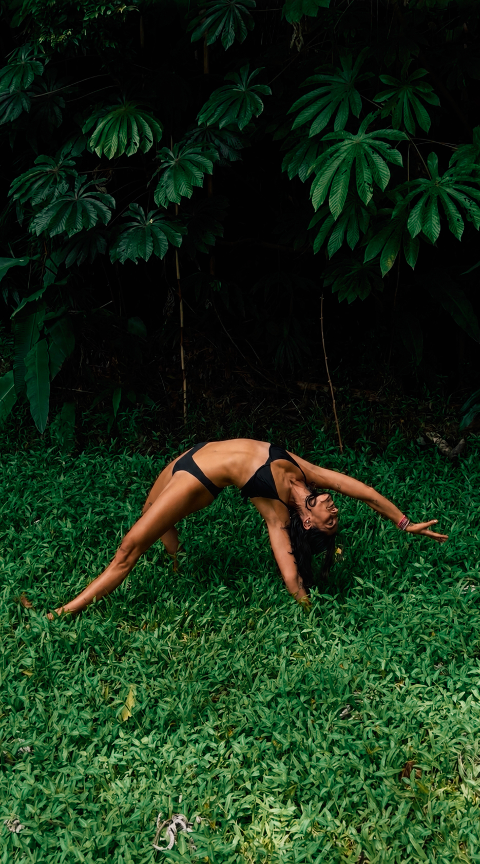 A woman in a backbend on a bed of vivid green jungle leaves — wild, embodied, tropical.
